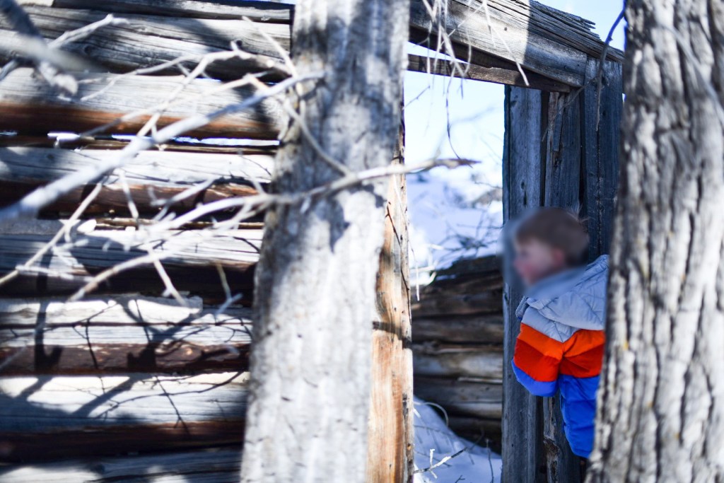 A child in colorful winter clothes peeks through the doorway of an old wooden cabin, surrounded by bare trees and snow.