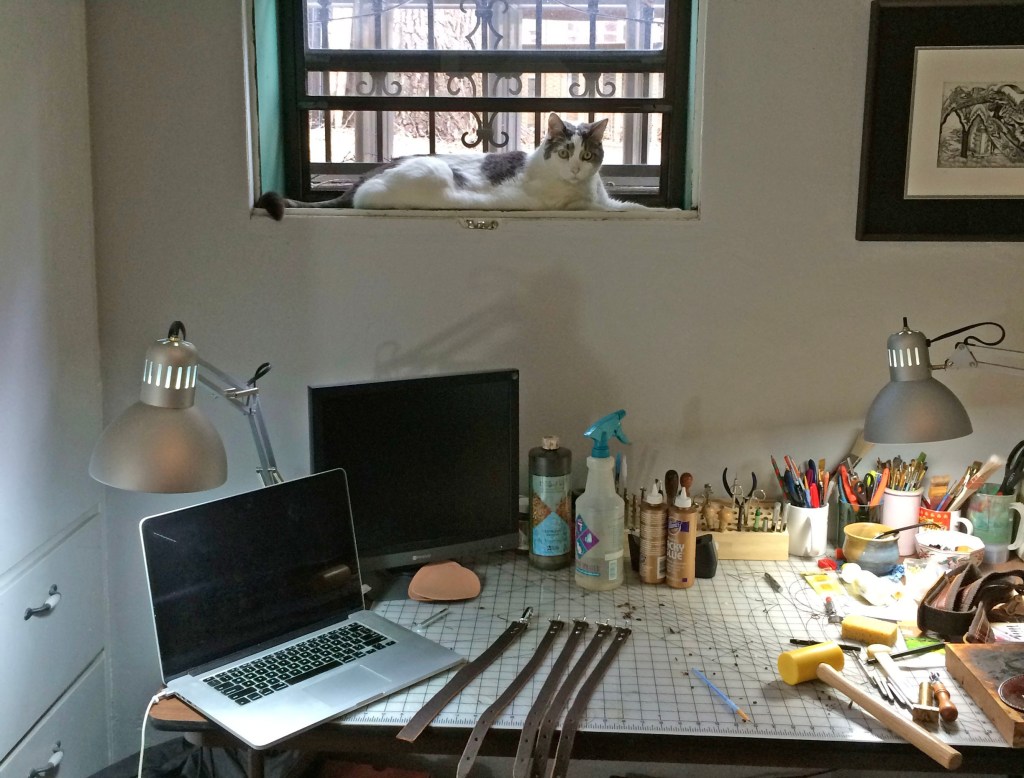 A workspace featuring a desk cluttered with leatherworking tools and materials, including cords, a laptop, and several containers. A cat is lounging on the windowsill in the background.